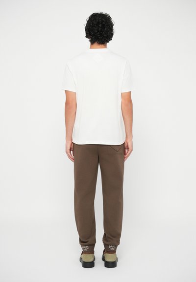 Man with curly hair wearing a white short-sleeve t-shirt, brown pants with back pocket, and brown and beige shoes, standing facing away.