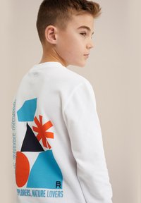 Fehér pulóver színes geometriai nyomtatással kék, narancs és fekete színben, a hátán "Felfedezők, Természetkedvelők" felirattal.