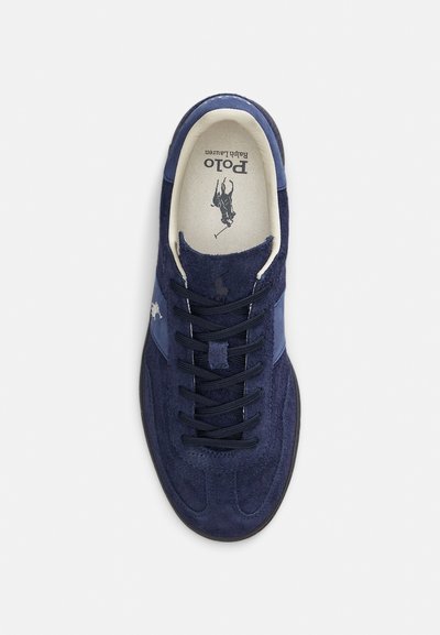 Sapatilha de camurça azul-marinho com um painel lateral azul-claro, atacadores pretos e interior bege com um logótipo impresso.