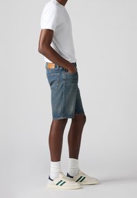 Mörkblå denim shorts för män med en urtvättad effekt, femficksdesign och en brun läderpatch på midjan, kombinerade med en vit t-shirt med rund halsringning.