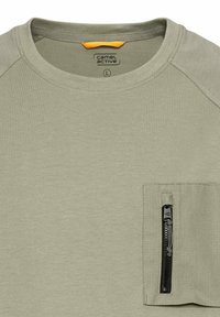 Kortærmet, lyseolivgrønt t-shirt lavet af blødt stof. Har en brystlomme med lynlås i sort og teksturerede stofdetaljer.