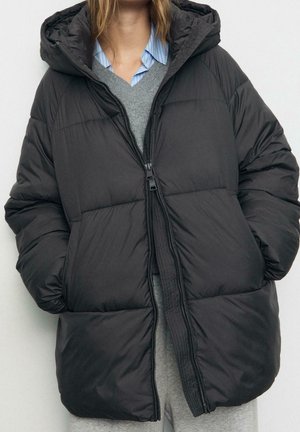 Femme portant une doudoune noire surdimensionnée avec capuche, mains dans les poches, par-dessus un pull gris et une chemise à col rayée bleue.