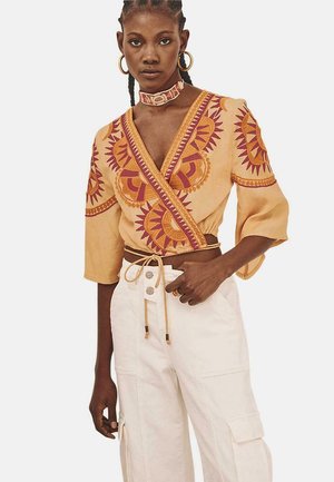 Blusa de estilo envolvente en tejido naranja claro con intrincados motivos de sol en rojo y morado, combinada con pantalones cargo blancos de cintura alta.