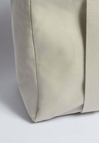 Plátěná taška v světle béžové barvě, se strukturovaným povrchem a pevnými tkanými popruhy. Design je jednoduchý a funkční s čistými liniemi.