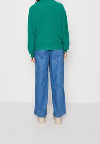 Grön stickad tröja med sänkt axel och ribbad nederkant, i kombination med löst sittande ljusblå denimbukser. Enfärgad bakgrund.