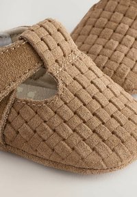 Chaussures en daim marron avec une texture tissée, présentant une forme arrondie et un côté ouvert. Détails de couture visibles le long des bords.