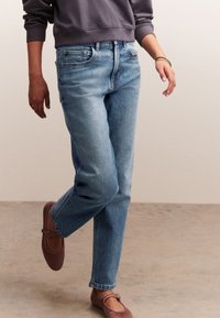 Világoskék farmernadrág, laza szabással és egyenes szárakkal. Barna lapos cipővel párosítva. Alap pulóver ujja látható fölötte.