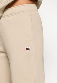 Φαρδύ μπεζ παντελόνι φούτερ με ομαλή υφή, που διαθέτει ένα μικρό μαύρο και κόκκινο κεντημένο λογότυπο στο αριστερό μηρό και ραφές στα πλάγια.