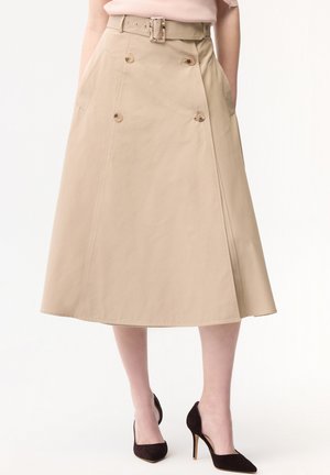 Beżowa spódnica midi w kształcie litery A z paskiem i guzikami, noszona z jasnoróżową bluzką i czarnymi szpilkami. Ręce w kieszeniach.
