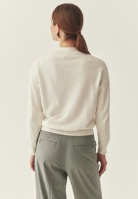 Pull tricot couleur crème à encolure arrondie et poignets côtelés, associé à un pantalon gris sur mesure avec poches arrière.