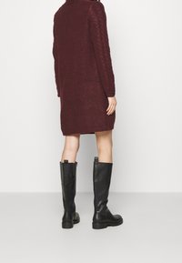 Bordowa dzianinowa sukienka z długimi rękawami, z teksturą w warkocze na ramionach. Połączona z czarnymi skórzanymi botkami sięgającymi do kolan.