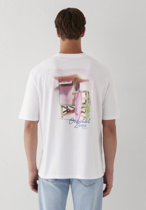 Mann trägt ein weißes T-Shirt mit bunter Rettungssessel- und Surfbrett-Grafik und Text „Coastal Zone“ auf dem Rücken, hellblaue Jeans.