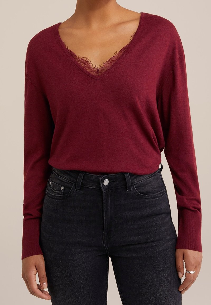 Femme portant un pull bordeaux à col en V avec bordure en dentelle, associé à un jean noir taille haute et une bague argentée à la main droite.