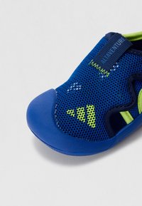 Blue mesh shoe features a textured rubber toe cap and green accents. Design includes cut-out sections for breathability and flexibility.