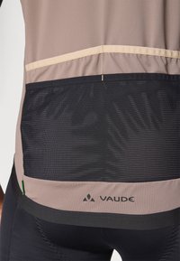 Vīriešu velo krekls brūnā krāsā ar melnu acs paneli aizmugurē, izceļoties ar brūnām šuvēm un VAUDE logo. Gluda tekstūra, sportiska piegriezuma.