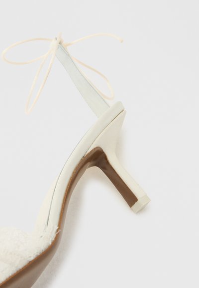 Λευκό πέδιλο με ψηλό τακούνι, λουράκι που δένει στον αστράγαλο, υφή στην περιοχή των δαχτύλων και λεπτό τακούνι σε απλό φωτεινό φόντο.