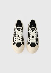 Tênis de lona pretos com detalhes brancos, apresentando atacadores e uma sola de borracha bege espessa. Detalhe do logótipo notável na lateral.