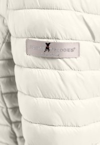 Biała kurtka puchowa o pikowanej teksturze. Posiada metkę z materiału z napisem "Frieda Freddies" oraz wyszywany czarny logo psa.