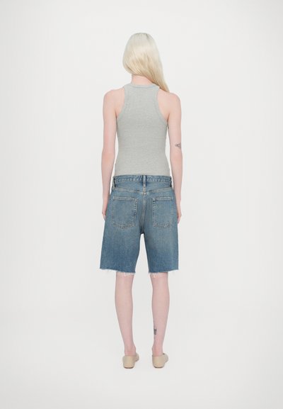 Hallitusest tank top, sinised denim lühikesed püksid purustatud äärega, kaks tagumist taskut ja beežid libisedavad kingad. Nähtav tätoveering paremal jalal.