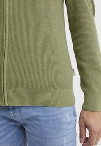 Olivenfarget strikket cardigan med hel glidelås, ribbestrikkede mansjetter og nederkant, og et lite merke på siden. Kombinert med lyseblå jeans.