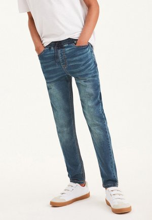 Dieťa v vybledlých modrých denimových teplákoch a bielych teniskách so suchými zipsami, ruky v nohaviciach, oblečené v bielom tričku.