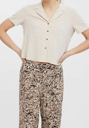 Vrouw draagt een beige kort mouwen overhemd met knopen, lichtjes ingestopt in een broek met hoge taille met zwarte en beige abstracte patronen.