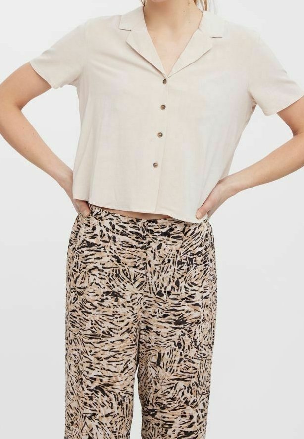 Femme portant une chemise beige à manches courtes boutonnée, légèrement rentrée dans un pantalon taille haute avec des motifs abstraits noirs et beiges.
