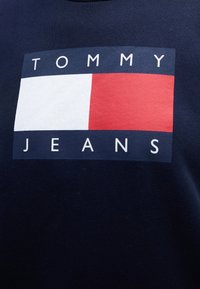 Tissu bleu marine avec un logo rectangulaire comportant des blocs blancs et rouges, ainsi que les mots « TOMMY » au-dessus et « JEANS » en dessous des blocs.