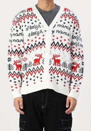 Hvid knap kardigan med røde og sorte zigzagmønstre, rensdyr og korsmønstre båret over en grå skjorte og sorte bukser.