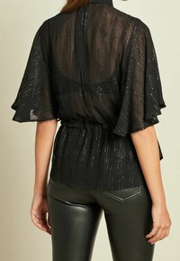 Blouse noire transparente avec manches volantées, taille froncée et accents en fil métallique. Associée à un pantalon ajusté noir effet cuir.