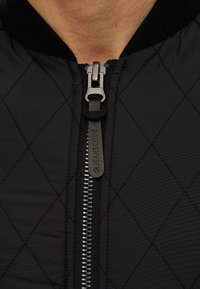 Czarna pikowana kurtka z wyraźnym zapięciem na zamek z teksturowanym uchwytem. Tkanina ma subtelny połysk i jest lekka.