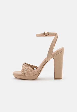 Beige højhælet sandal med flettede forreste stropper, ankremed guldbøjle og en chunky blokhæl på hvid baggrund.
