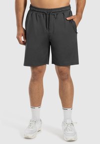 Graue Sportshorts mit elastischem Bund und Kordelzug. Ausgestattet mit Seitentaschen und einer lockeren Passform. Getragen mit weißen Sneakers und Socken.