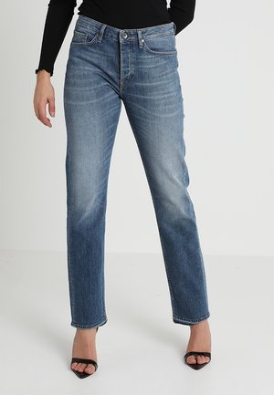 Frau trägt blaujeans mit mittlerer Taille und geradem Bein, kombiniert mit schwarzen offenen Sandalen mit Absatz und einem schwarzen Langarmshirt.