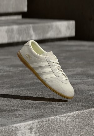 Biały zamszowy sneaker z trzema teksturowanymi białymi paskami, gumową podeszwą w kolorze brązowym oraz złotym logo po boku, cechujący się minimalistycznym designem.