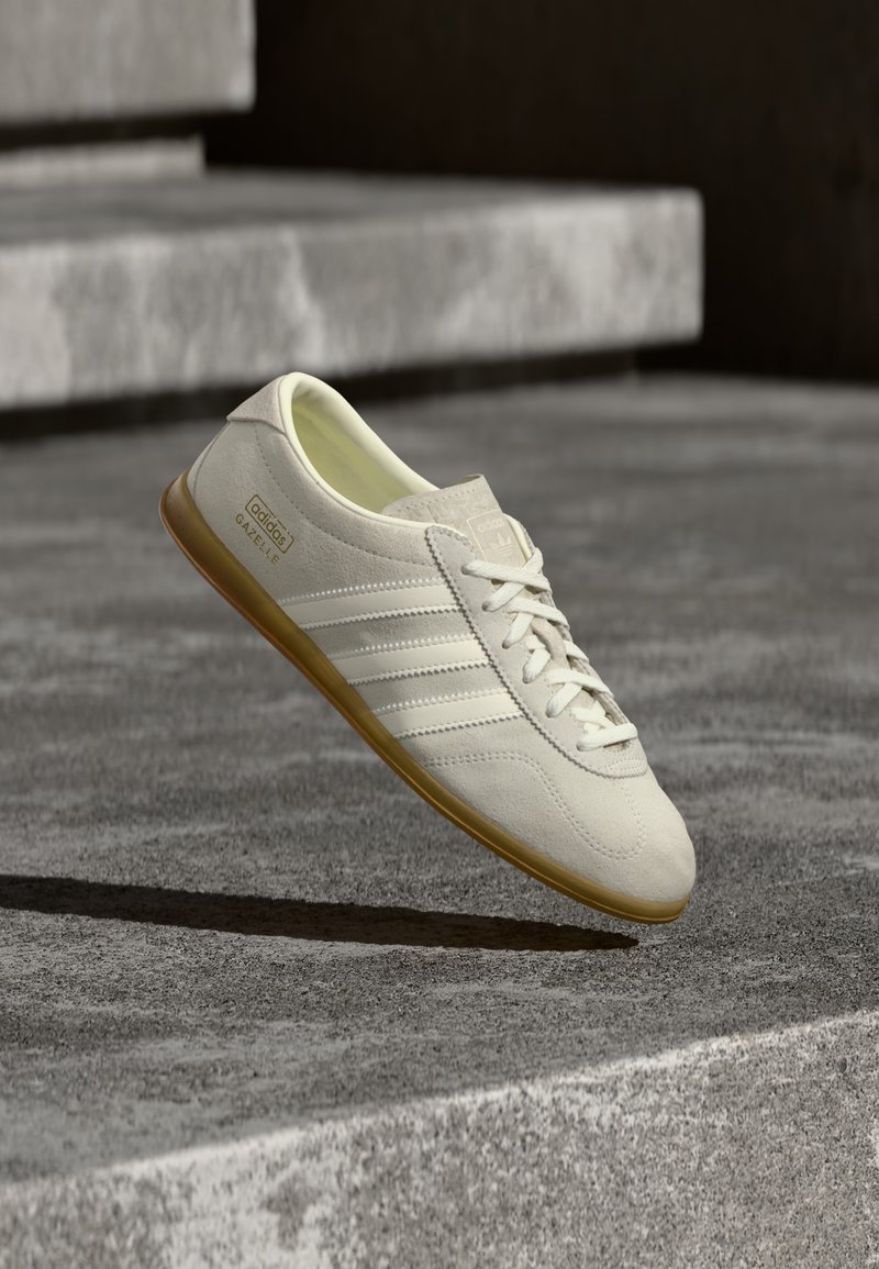 Biały zamszowy sneaker z trzema teksturowanymi białymi paskami, gumową podeszwą w kolorze brązowym oraz złotym logo po boku, cechujący się minimalistycznym designem.