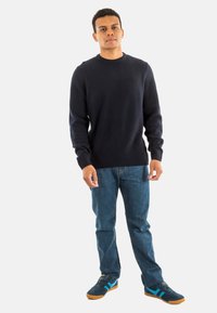 Marine crew-neck trui van zachte stof, gecombineerd met blauwe denim jeans en donkere sneakers met turquoise accenten. Alleenstaande pose, neutrale achtergrond.