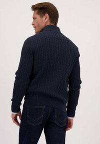 Man die met zijn rug naar de camera staat en een marineblauwe kabelgebreide trui over een shirt draagt, samen met donkerblauwe jeans met contrasterende stiksels op de zakken.