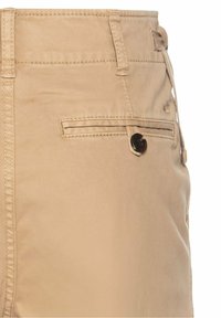 Chino beige avec une poche arrière boutonnée, des passants de ceinture et des détails de couture visibles sur un fond uni.