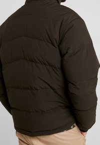 Brun pufferjakke med kviltet design, højt krave og elastiske manchetter. Stoffet ser glat ud med en mat finish.