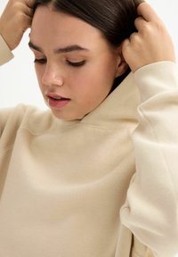 Sudadera beige con capucha de ajuste holgado, textura suave, puños acanalados y costuras decorativas en los hombros.