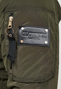 Chaqueta verde oliva con un bolsillo con cremallera y herrajes dorados, un parche de cuero negro con texto en relieve y tela texturizada.