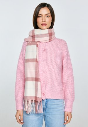 Różowy sweter na guziki, zestawiony z różowo-beżowym szalikiem w kratę z frędzlami na końcach. Dopełnieniem stroju są jasnoniebieskie dżinsy.