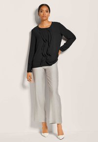 Zwarte blouse met lange mouwen en verticale volants, gecombineerd met glanzende lichtgrijze wijde broek en witte spitse hakken.