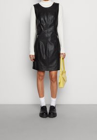 Schwarzes Lederkleid mit figurbetontem Schnitt, ausgestattet mit Reißverschlusstaschen, kombiniert mit einem weißen Rollkragenpullover, einer gelben Handtasche und schwarzen Schuhen.