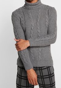 Personne portant un pull à col roulé gris en maille torsadée et un pantalon à carreaux gris, avec un bras croisé sur l'autre, sur un fond uni.