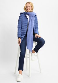 Niebieska, ocieplana kurtka z kapturem i zamkiem błyskawicznym, w połączeniu z jasnoniebieskim, miękkim szalikiem. Model nosi ciemne dżinsy i białe trampki, siedząc na stołku.