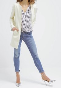 Vit skräddarsydd kappa med långa ärmar, blus med prickar, ljusblå smala jeans och silverfärgade högklackade skor. Smidiga texturer och stilren design.