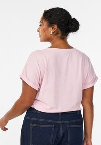 Camiseta rosa claro con mangas remangadas, tela suave, corte relajado y cuello redondeado, combinada con vaqueros de mezclilla oscuros.