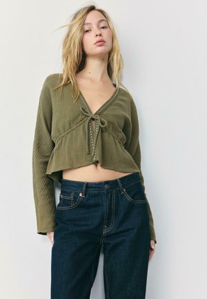 Junge Frau mit langen blonden Haaren, die ein olivgrünes, langärmliges, kurz geschnittenes Oberteil mit Knoten vorne und dunkelblaue, hoch taillierte Jeans trägt.